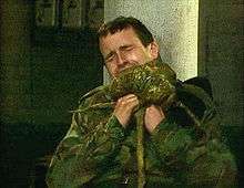 A man in camouflage fatigues winces with pain as he tries to remove a green alien creature from his neck.