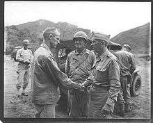 Two men in uniform shake hands. One is bear headed; Krueger wears his garrison cap. With them is a man wearing a steel helmet. A jeep is parked behind them.