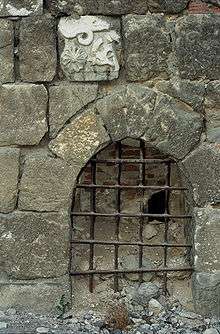 A small gate with iron bars set in a wall built of uneven stones, one of which is decorated