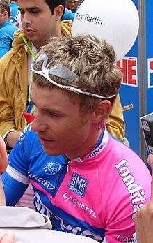 A man signing autographs while wearing a pink and blue cycling jersey.