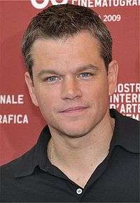 A head shot of Matt Damon, a caucasian male in his late-30s with dark hair, looking into the camera smiling slightly. He wears a black polo shirt, and stands in front of a red background with white font.