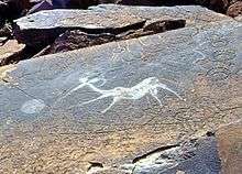 A sandstone slab in horizontal position covered by a dark patina that appears greasy black as if dirtied by touch. In the middle of the slab is a clear white indentation of a fantasy creature with the body of an antelope and the torso and head of a human who appears to b shooting at something with a bow. The figure is surrounded by other engravings that appear geometric and have the color of the slab surface.