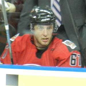 An ice hockey player sitting on the bench. He is wearing a black helmet with a visor and a red jersey with the number 61 on the sleeves.