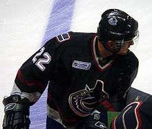 An ice hockey player leaning over on his bench with his head turned to the right. He wears a dark blue jersey and a visored helmet.
