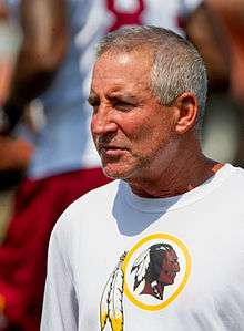 Candid chest-up photograph of Smith with close-cropped grey hair wearing a white t-shirt with a Washington Redskins logo