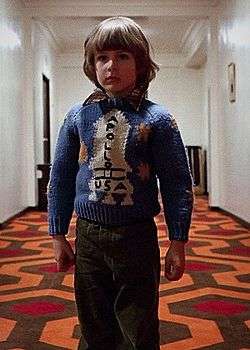 Danny Lloyd as the child character Danny Torrance. He stands in a hotel hallway and wears a sweater with a rocketship on it.