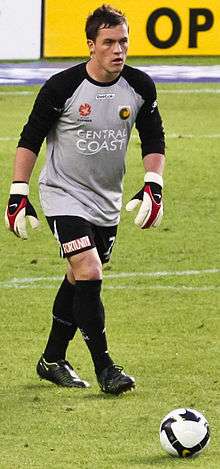 A man with dark hair, wearing a grey shirt, black shorts and goalkeeping gloves approaching a football