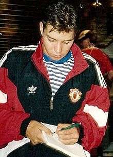 A photograph of a man with short brown hair wearing a black tracksuit top with red trim over a blue and white hooped shirt. He is signing an autograph.
