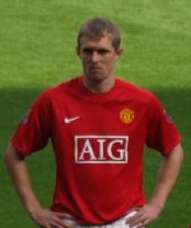 A white man with light hair standing with his hands on his hips. He is wearing a red football shirt.