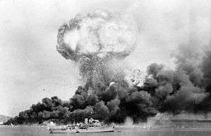 Long shot of mushroom cloud from an explosion, and black billowing smoke from nearby fire, with ship in foreground