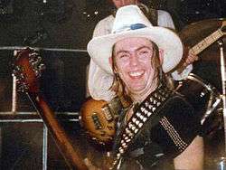 A man with shoulder-length hair wears a black t-shirt and a white cowboy hat.  His guitar sling sports studs.