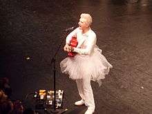 A bird's-eye view of David Byrne playing acoustic guitar and singing into a microphone onstage while wearing a white jumpsuit and tutu