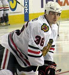 Hockey player in white uniform with a picture of crossed hockey sticks on the shoulder and the painted face of an indigenous person on the chest. He is crouched on the ice, feet further than shoulder width apart.