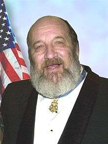 Head and shoulders of a bearded white man wearing a black dress jacket with white shirt and a medal hanging from his neck.