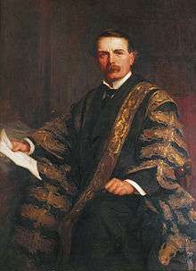 A man in his late 40s, with brown hair and moustache, wearing a black suit, white wing-collared shirt and black tie, together with a black gown repeatedly patterned in gold brocade; he sits in a chair, holding a piece of paper in his right hand