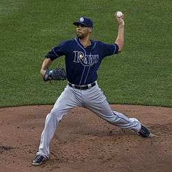 A man in gray pants, a blue baseball jersey with 'Rays' on the chest, and a blue baseball cap is in the process of pitching a baseball with his left hand.