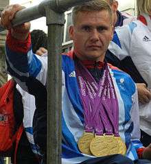A man in a wheelchair, he wears a white and blue top and around his neck hang four gold medals.