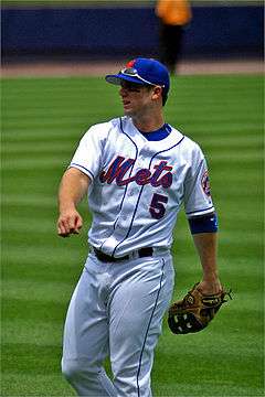 Man in a white baseball uniform with "Mets" and "5" on the chest wearing a baseball glove on his left hand and a blue baseball cap.