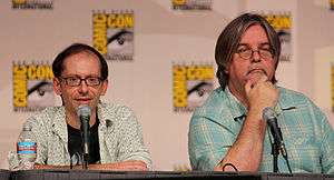 Two men sit at a table behind microphones, both have glasses, and one is shorter than the other.