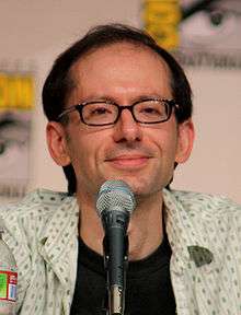 A closeup of a man in front of a microphone. He has a receding hairline and wears dark-framed glasses.