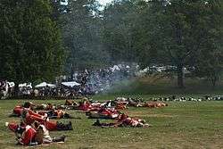 An open grassy area is strewn with bodies, most of them in red and white uniforms, although some with blue coats are visible farther back. In the distance there are white tents and a crowd of people, and a small thin cloud of smoke obscures the view a little.