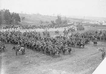 A large group of men and horses drawn up into lines in a field and on the adjoining road. A hill with trees and tents can be seen in the background.