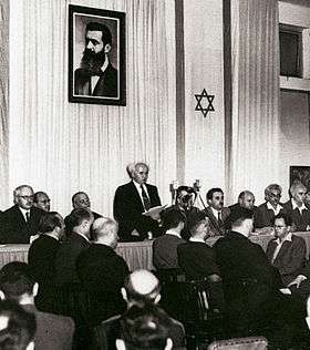 A single man, adorned on both sides by a dozen sitting men, reads a document to a small audience assembled before him. Behind him are two elongated flags bearing the Star of David and portrait of a bearded man in his forties.