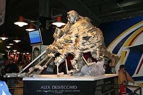 A large bronze statue of an ice hockey player in the act of shooting the puck.  The player's hands and stick are shown numerous times in order to simulate motion. At the base of the statue is inscribed "Alex Delvecchio Born: December 4, 1931 Fort William, Ontario, Canada