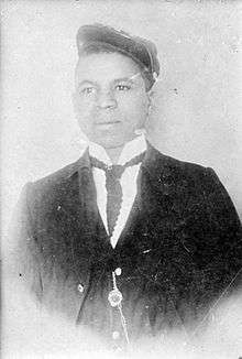 Head and torso of a young black man wearing a suit and tie with a watch chain hanging from a jacket button. He has a cap pushed high up on his forehead and tilted over his left ear.
