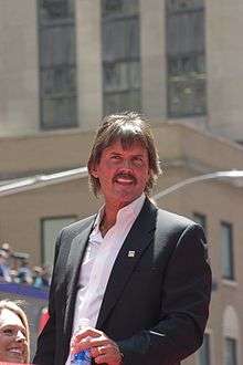 Dennis Eckersley in a black suit with the top of a water bottle in his right hand