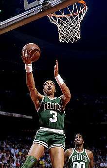 A man, wearing a green jersey with a word "CELTICS" and the number "3" written in the front, is jumping while holding a basketball, trying to do a layup.