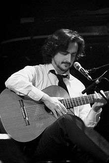 Black-and-white picture of a man in his early 30s. He has long hair and beard and moustahce, and wears a shirt, a tie, and trousers. He is sitting with an acoustic guitar and a microphone by his face.