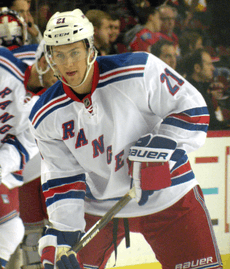 An ice hockey player leaning forward with his ice hockey stick resting on the ice. He is wearing a white helmet and a red, white and blue uniform.