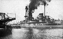 A large warship sits in harbor, smoke pouring from its funnels.