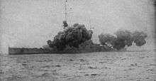 A large warship is partially obscured by smoke from its main guns firing
