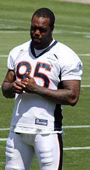 Color photo of African-American football player Derrick Harvey in the white, navy blue and orange uniform of the Denver Broncos, standing on the side of a playing field with his helmet off.