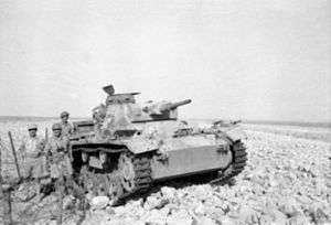 A tank lies crippled in the desert with soldiers standing around it