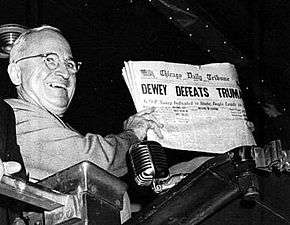 Man in gray suit and wire glasses holding newspaper that says "Dewey Defeats Truman"