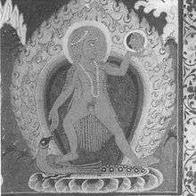 A nude woman with long hair and wearing a pearl necklace and headband, stands on a peacock (which in turn sits on a lotus) with her legs apart. She holds a mirror in her left hand and looks at her reflection in it. An aureole and halo surround her.