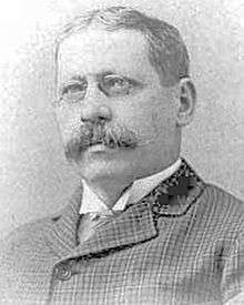 A black and white photograph of the head and shoulders of a man with a mustache and glasses