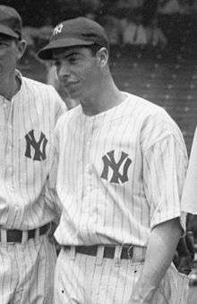 A man stands in a line. He is wearing a white uniform with stripes and a black baseball cap, which both have an interlocking "N" and "Y".