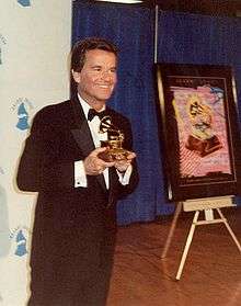 A smiling, formally clad man holding a Grammy Award.