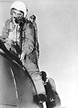 Mustachioed man in flying suit and helmet alighting from cockpit