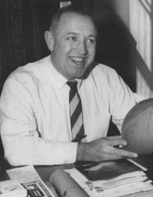 Dick Gallagher sitting at a table with a football in his hands in 1959