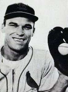 A smiling man in a baseball uniform with a cardinal on the left breast holds a baseball in a glove on his left hand.