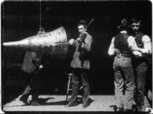 Black-and-white film screenshot of a man (at center) playing a violin while facing a large cone. An obscured man is seen behind the cone. Two men at the right are dancing with each other.