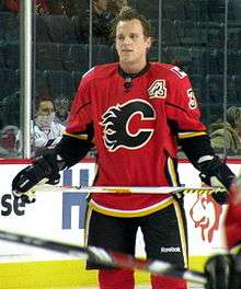 A Calgary Flames player observes his teammates who are off camera.  On his uniform is a small patch that uses Atlanta's "Flaming A" logo to denote his position as an alternate captain.