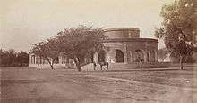 Old photo of low, round building with trees, a man and a horse in front