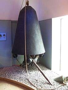 A photograph of a bell-shaped metal chamber with a metal platform suspended from its edges. The chamber is turned with the open end down. Inside stands a mannequin extending a long ship's hook as if searching for something to hook on to.