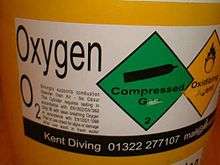  The white adhesive plastic label displays the gas name, Oxygen, and the chemical symbol O2 with a block of small text on the left side describing the hazards of the contents, then a green diamond symbol for compressed gas and a yellow diamond for oxidising agent.